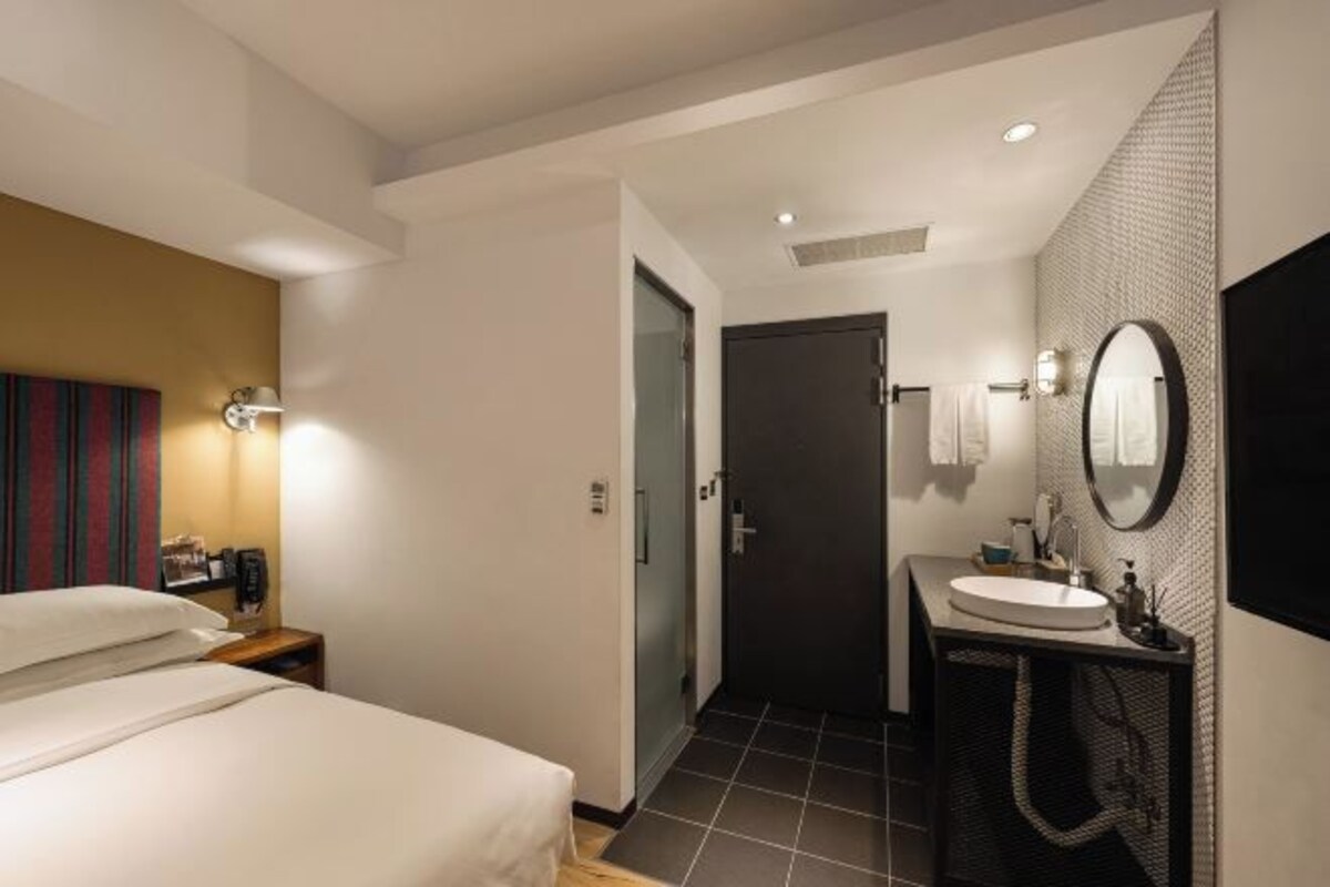 Una habitación de hotel con una cama, un baño con lavamanos y espejo, y una puerta que da a otra estancia.