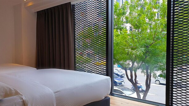 A modern hotel room with a large window, a bed with white linens, and a view of the street outside. - River inn Station (Kaohsiung)