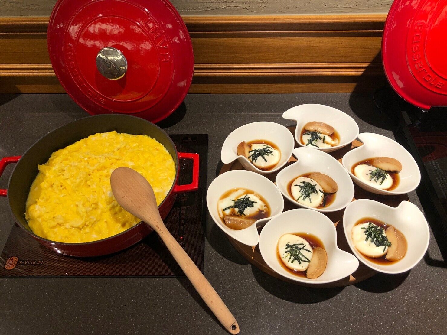 A kitchen counter with a red pot of scrambled eggs, a wooden spoon, and several small bowls with a white substance and green garnish.