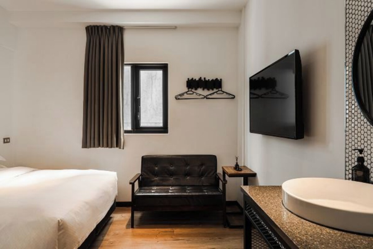 Habitación de hotel con cama, sofá de cuero, televisión, estante montado en la pared y baño con lavamanos y espejo.