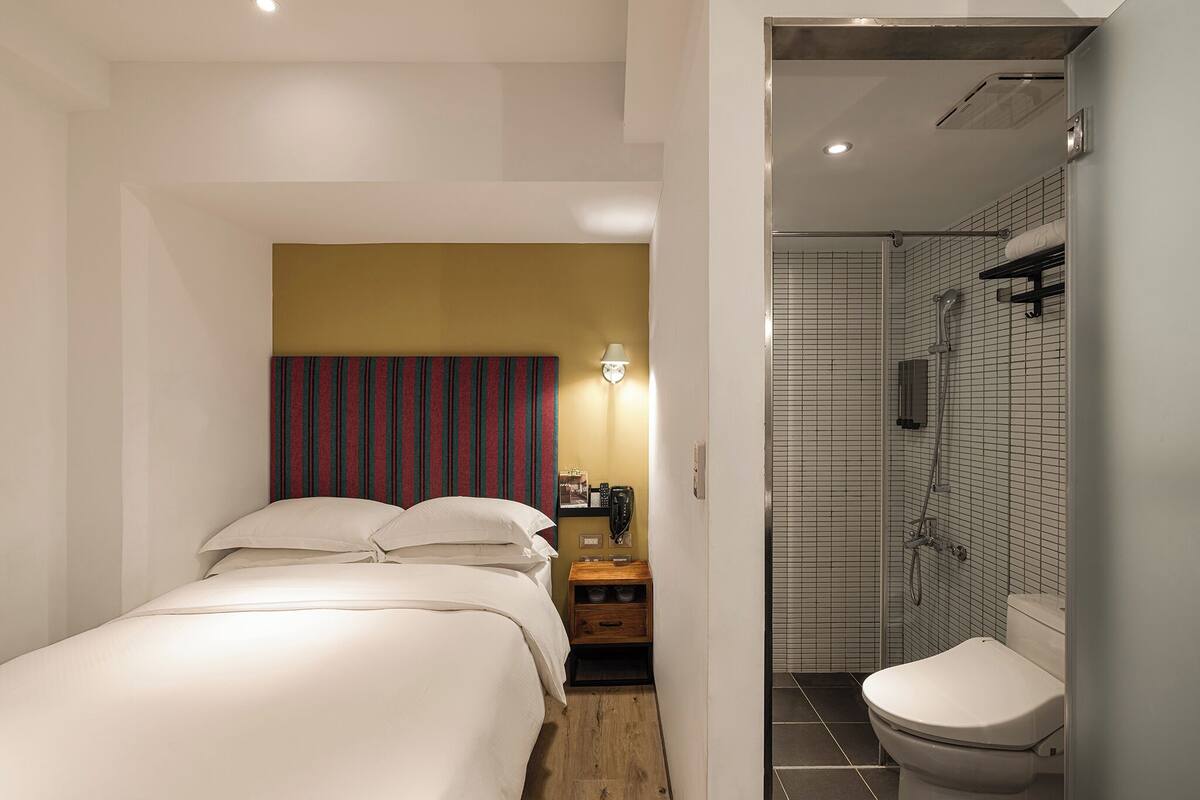 Quarto de hotel com cama, mesa de cabeceira, abajur e área de chuveiro com vaso sanitário.
