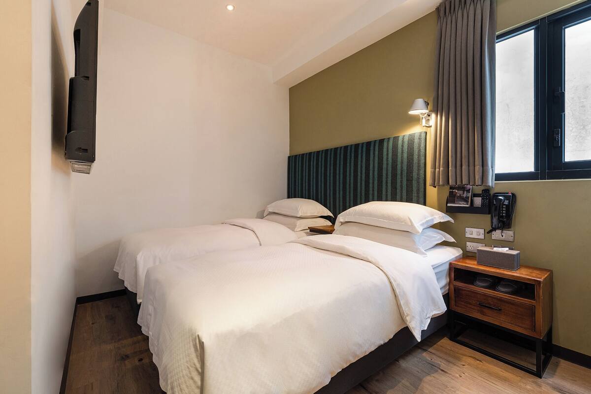 Quarto de hotel com cama, mesa de cabeceira, abajur e janela com cortinas.