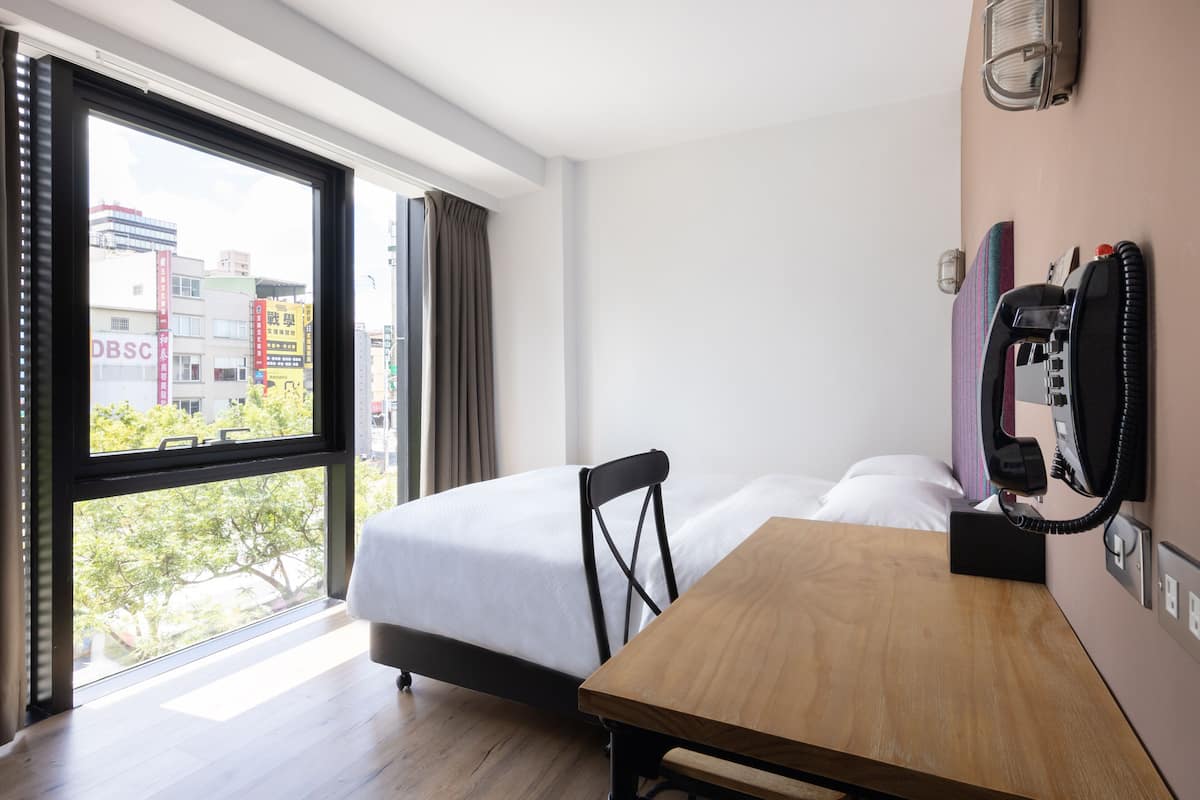 Quarto de hotel com cama, mesa de madeira, telefone e janela com vista para a cidade.
