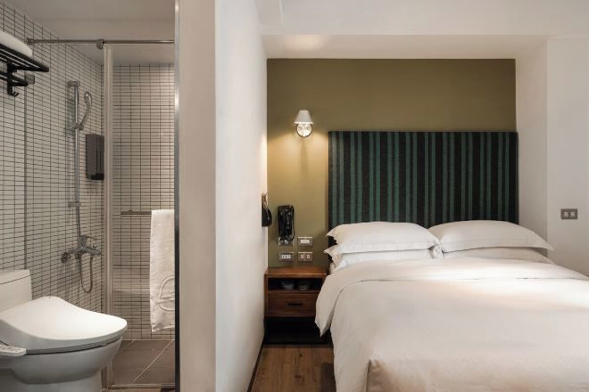 A hotel room with a shower, toilet, bed, bedside table, and a wall-mounted phone.