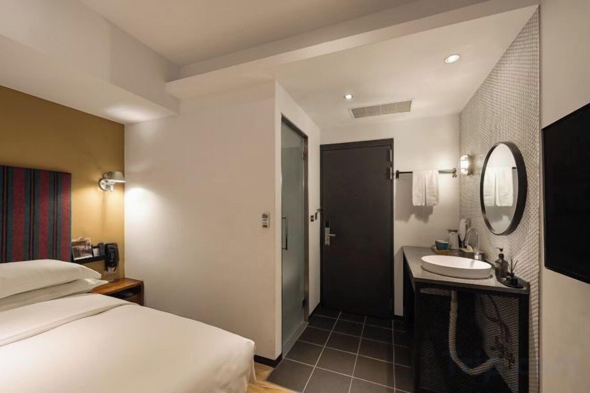 A hotel room with a bed, a bedside table, a bathroom with a sink and mirror, and a door leading to another room.