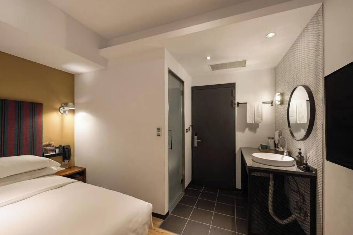 Quarto de hotel com cama, banheiro com pia e espelho, e porta que dá para outro cômodo.