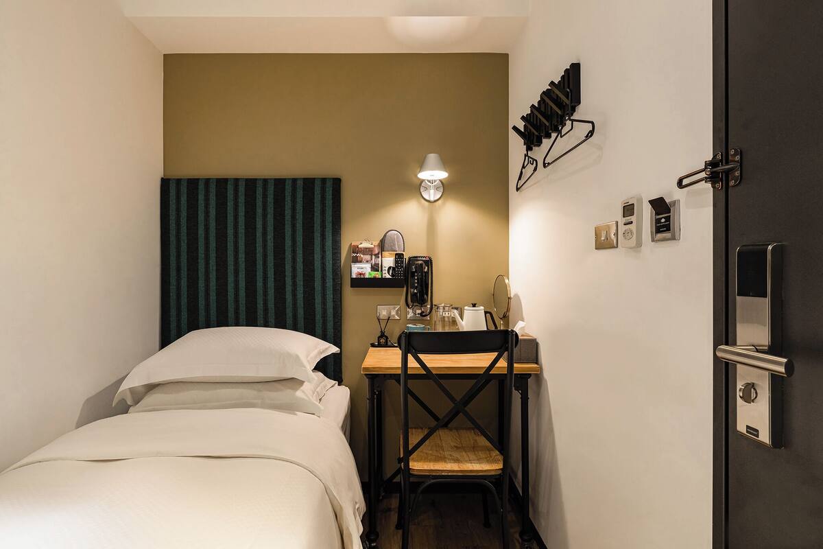 Quarto de hotel pequeno com cama, escrivaninha e cadeira, luminária e porta com fechadura.