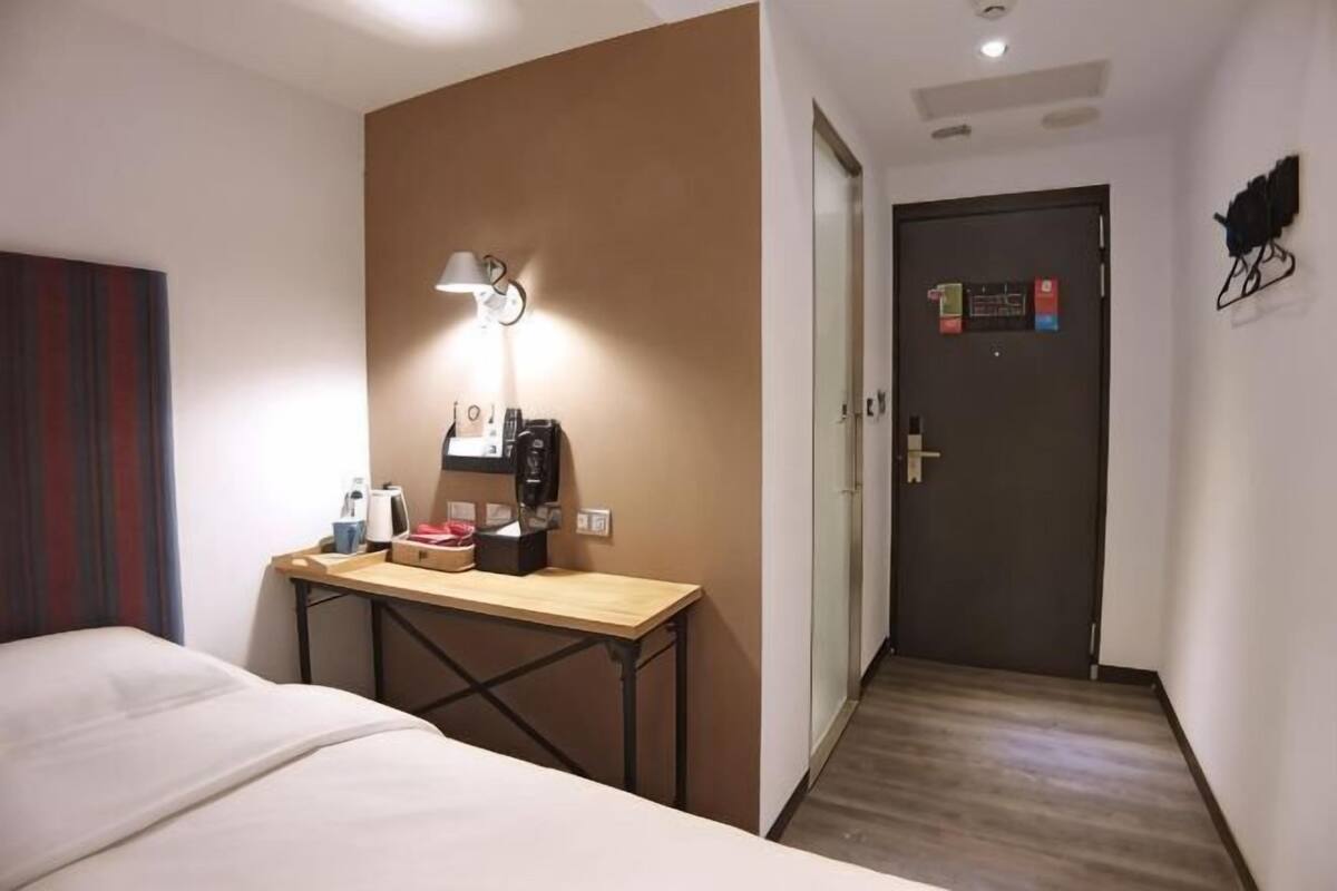 Quarto de hotel com uma cama, uma escrivaninha com luminária, uma cafeteira e uma porta com placa.
