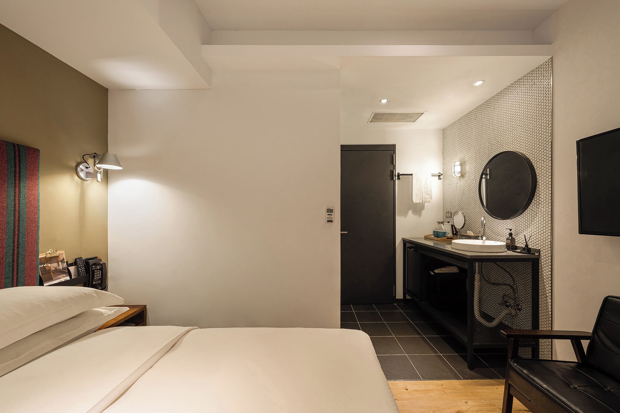 A hotel room with a bed, bedside table, lamp, and a view into the bathroom with a sink and mirror.