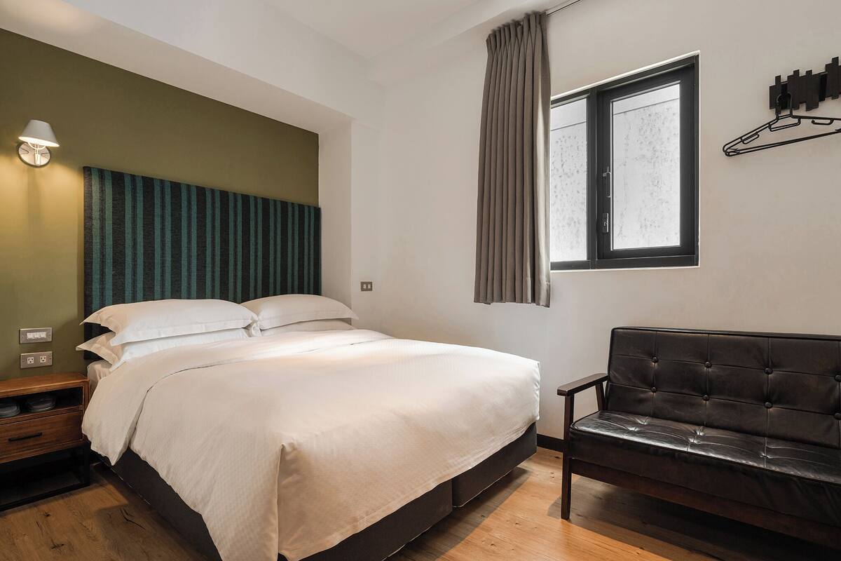 Quarto com cama, mesa de cabeceira, abajur, janela e um sofá.