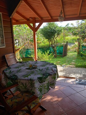 Un patio couvert avec une table et des chaises en bois, un hamac et une vue sur un jardin luxuriant.