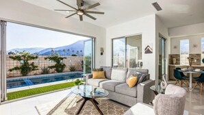A modern living room with a glass coffee table, a grey sofa with cushions, and a view of a swimming pool and mountains. - Mountain View Luxury Resort-Style Modern Home with a Private Pool. (Palm Springs)