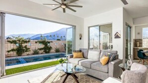A modern living room with a glass coffee table, a grey sofa with cushions, and a view of a swimming pool and mountains.