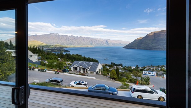 A view from a window overlooking a parking area with several cars, a building with a sloped roof, and a mountainous landscape with a body of water in the background.