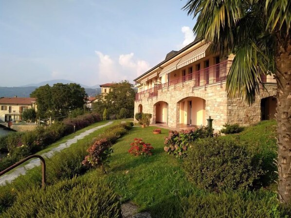 A stone building with a balcony, surrounded by lush greenery and a pathway.