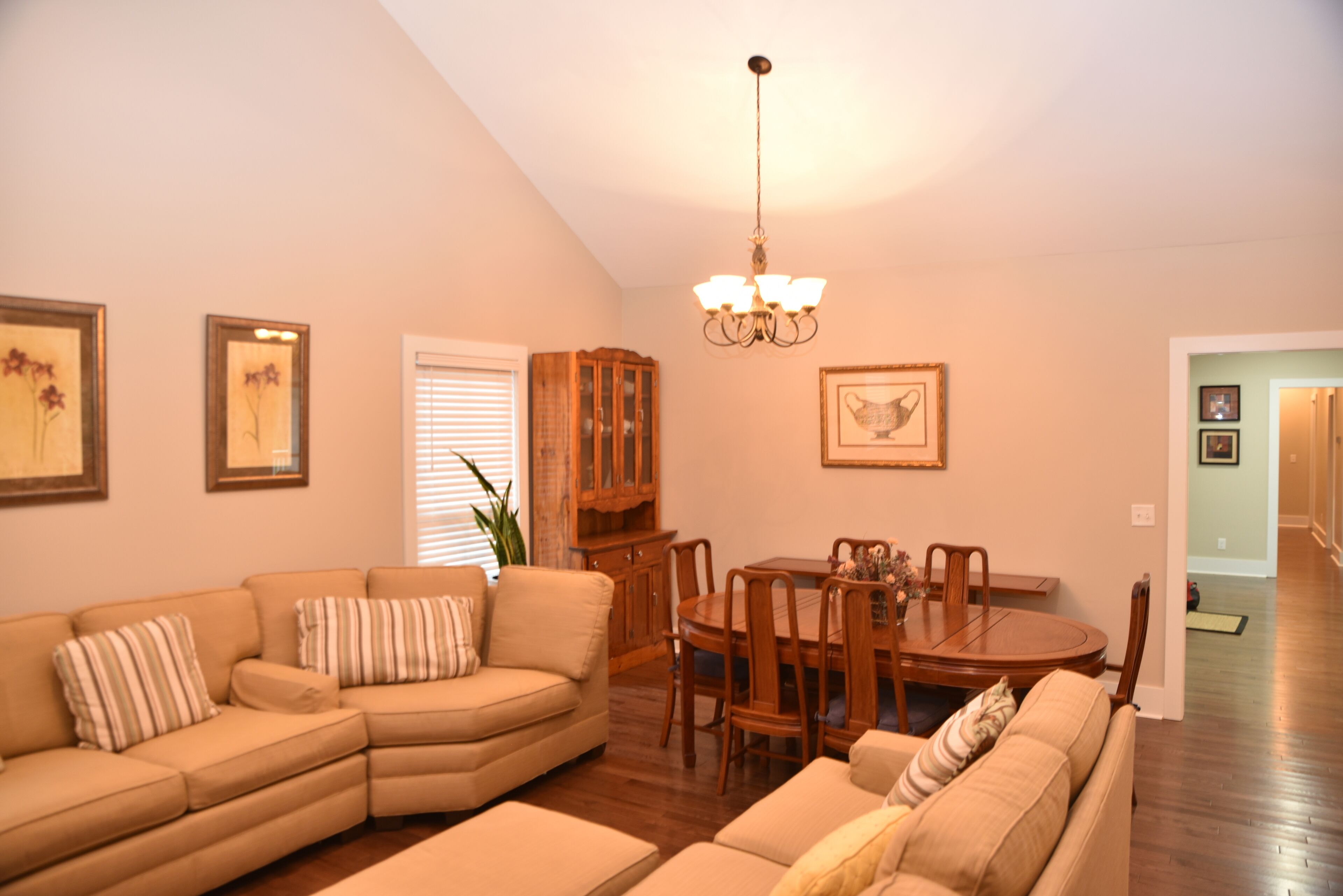 A living room with a sectional sofa, a dining table with chairs, a chandelier, and framed artwork on the walls.