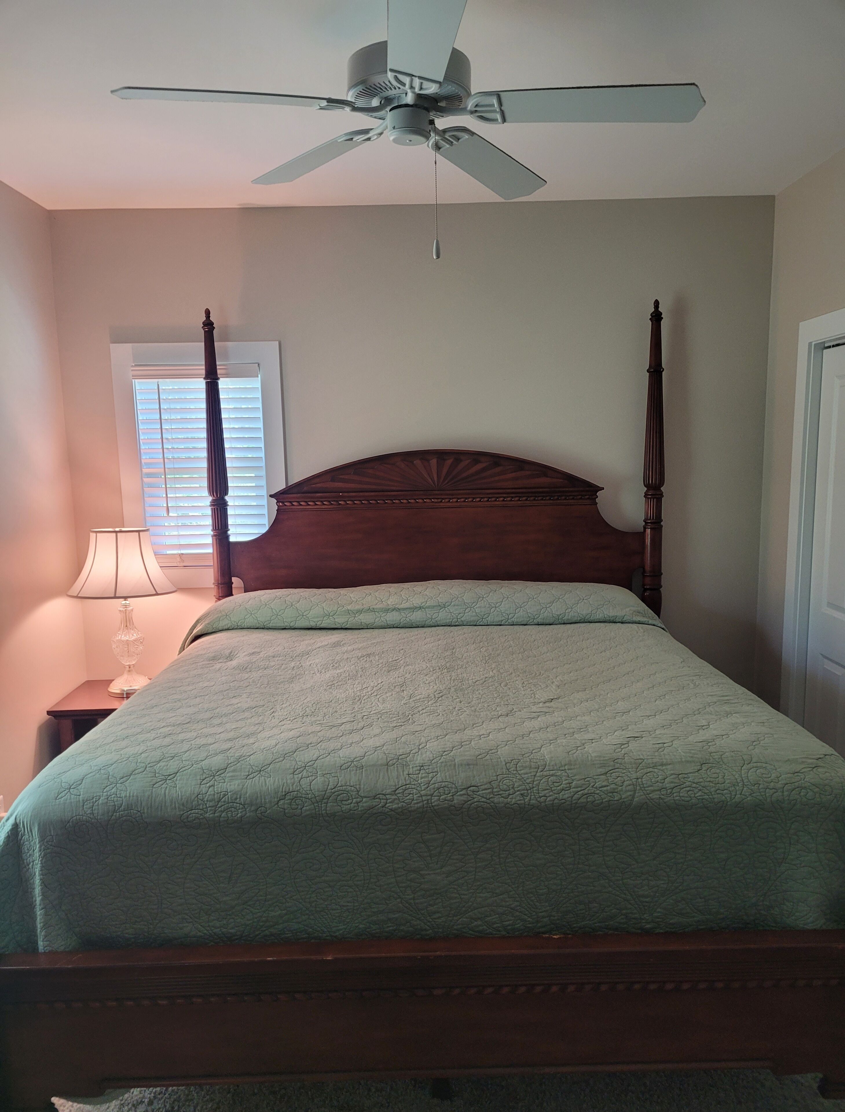 A bedroom with a four-poster bed, a ceiling fan, a bedside lamp, and a window with blinds.