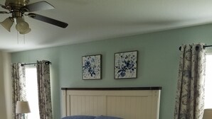 Un dormitorio con una cama, una mesita de noche, un ventilador de techo y dos cuadros con motivos florales.