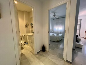 A hallway leading to a bathroom with a sink and mirror, and a bedroom with a bed and curtains. - Apartment Alextia | Apartelius | pool and WiFi (Chiclana de la Frontera)