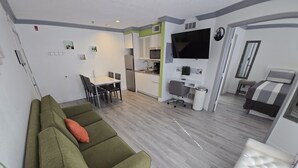 Uma sala de estar moderna com um sofá verde, uma cabeceira estofada branca, um espaço de jantar com mesa e cadeiras, uma cozinha com geladeira e micro-ondas, e uma TV fixada na parede.