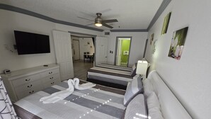 Quarto de hotel com uma cama grande, televisão de tela plana, ventoinha no teto e um banheiro visível através de uma porta aberta.