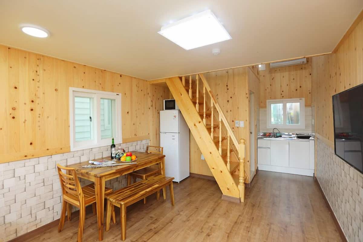 Una cocina compacta con paredes de madera, un comedor con una mesa y sillas de madera, un refrigerador blanco y una escalera de madera.