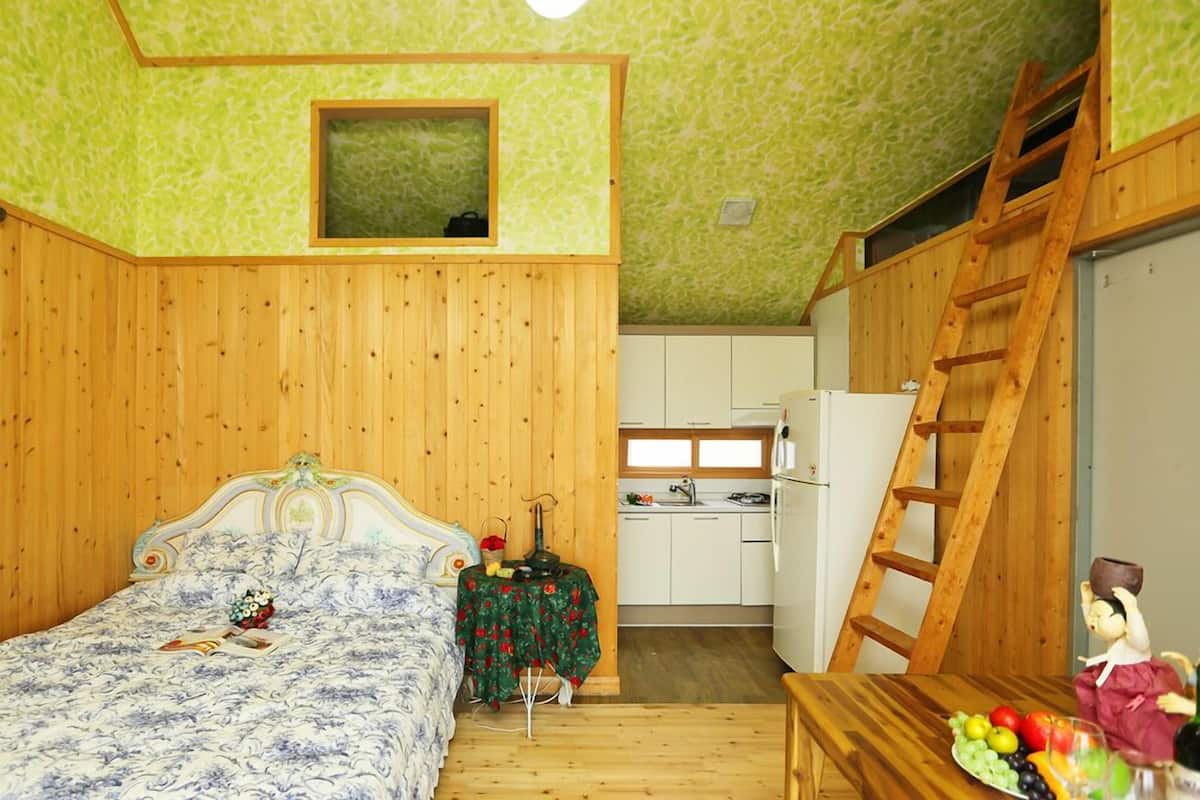 Una habitación pequeña y acogedora con una cama de madera, una mesita con una bandeja de fruta, una escalera y una kitchenette al fondo.