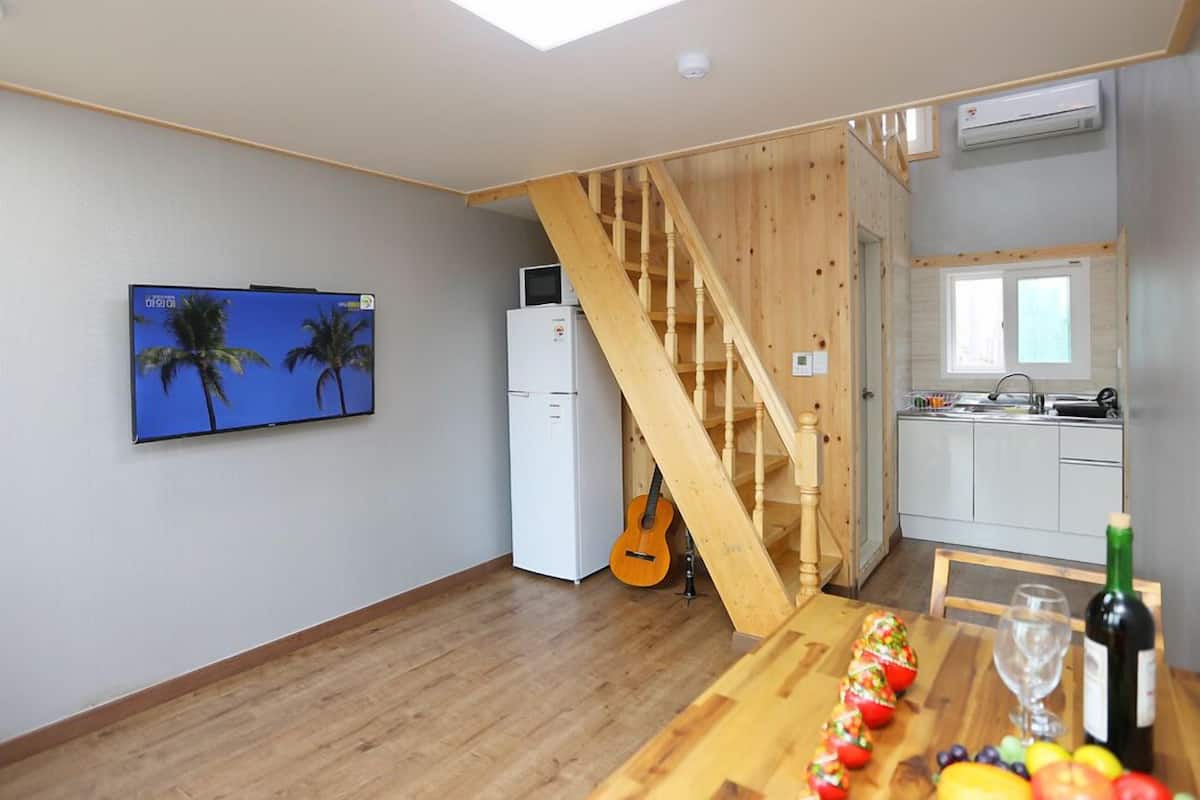 Una habitación con una escalera de madera, un televisor montado en la pared, un refrigerador, una zona de cocina con un fregadero y una mesa de comedor con frutas y una botella.