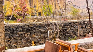 A stone wall with wooden benches and chairs, a view of traditional buildings, and a tree with autumn leaves. - BSR Farms (Pauri)