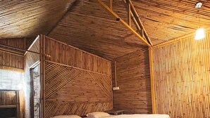 A room with a bed, a small table, and a chair. There is a wooden ceiling and walls, and a window on the left side. - BSR Farms (Pauri)