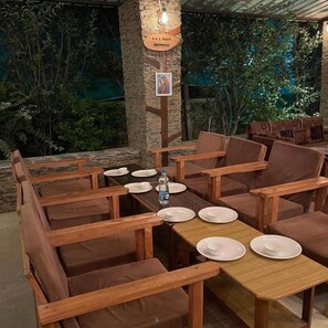 A restaurant with wooden tables and chairs, a sign with a picture, and a view of greenery. - BSR Farms (Pauri)