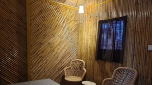 A room with a bed, two wicker chairs, a small round table, and a television mounted on the wall. - BSR Farms (Pauri)