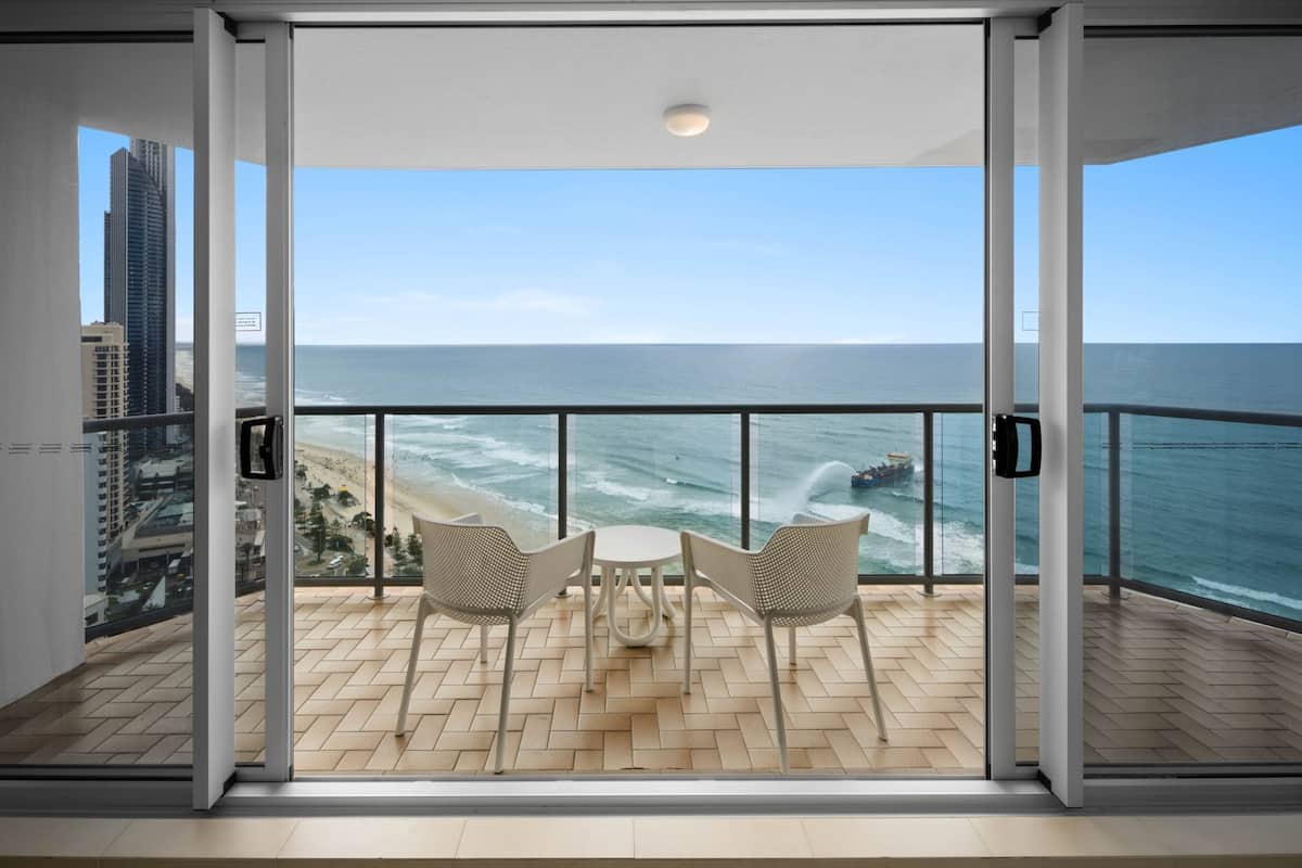 A balcony with a view of the beach and ocean, featuring two wicker chairs and a small table.