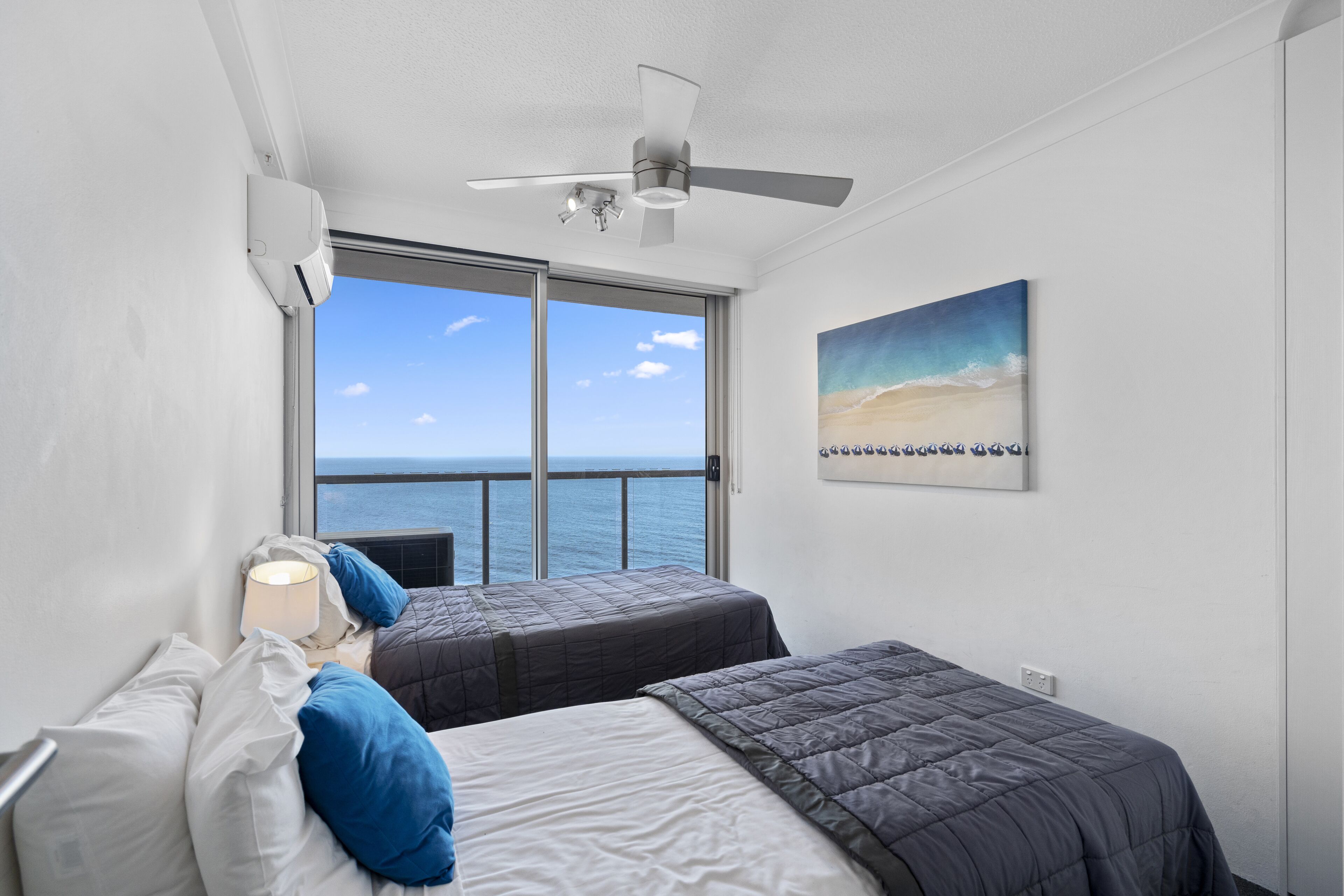 A hotel room with two beds, a ceiling fan, a large window with a view of the sea, and a painting on the wall.