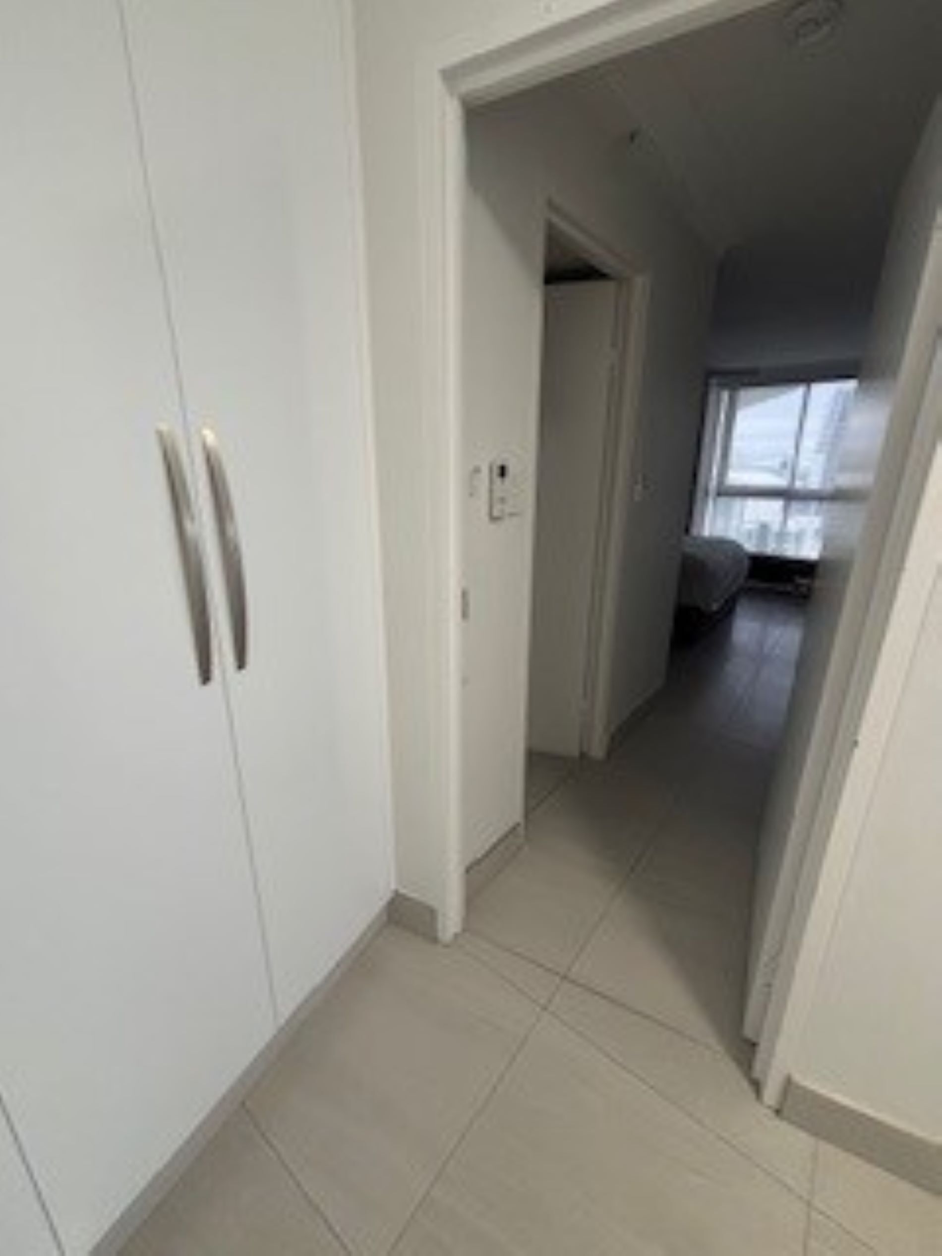 A hallway with white walls and a tiled floor leading to a room with a bed and a window.