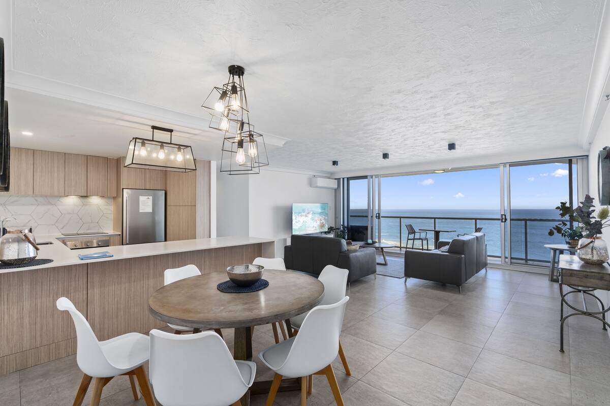 A modern kitchen with a large island, white chairs, and a dining table. There is a view of the ocean through large windows.