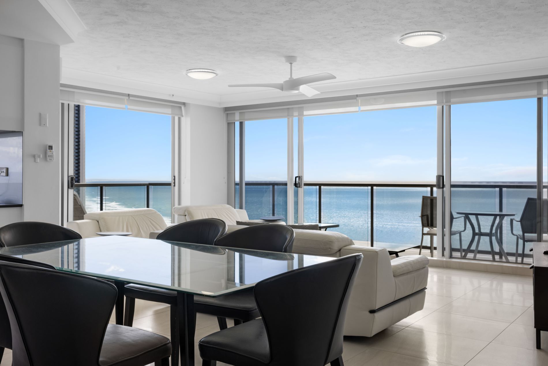 Una habitación moderna con una mesa y sillas de vidrio, ventanas amplias con vista al mar y un ventilador de techo.