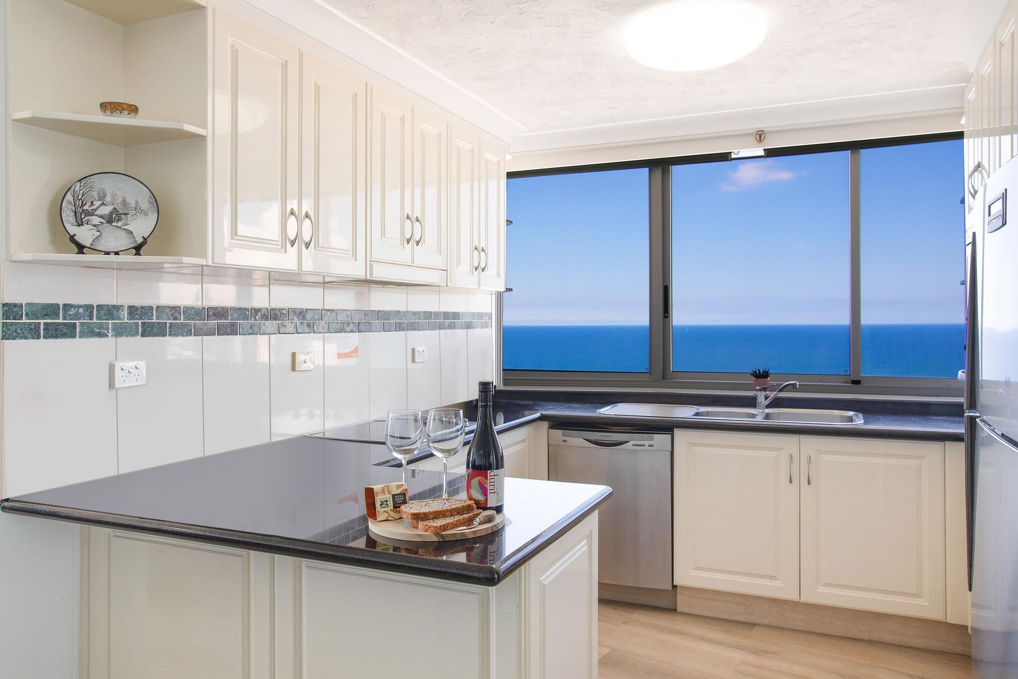 A modern kitchen with white cabinets, a black countertop, and a large window offering a view of the sea.