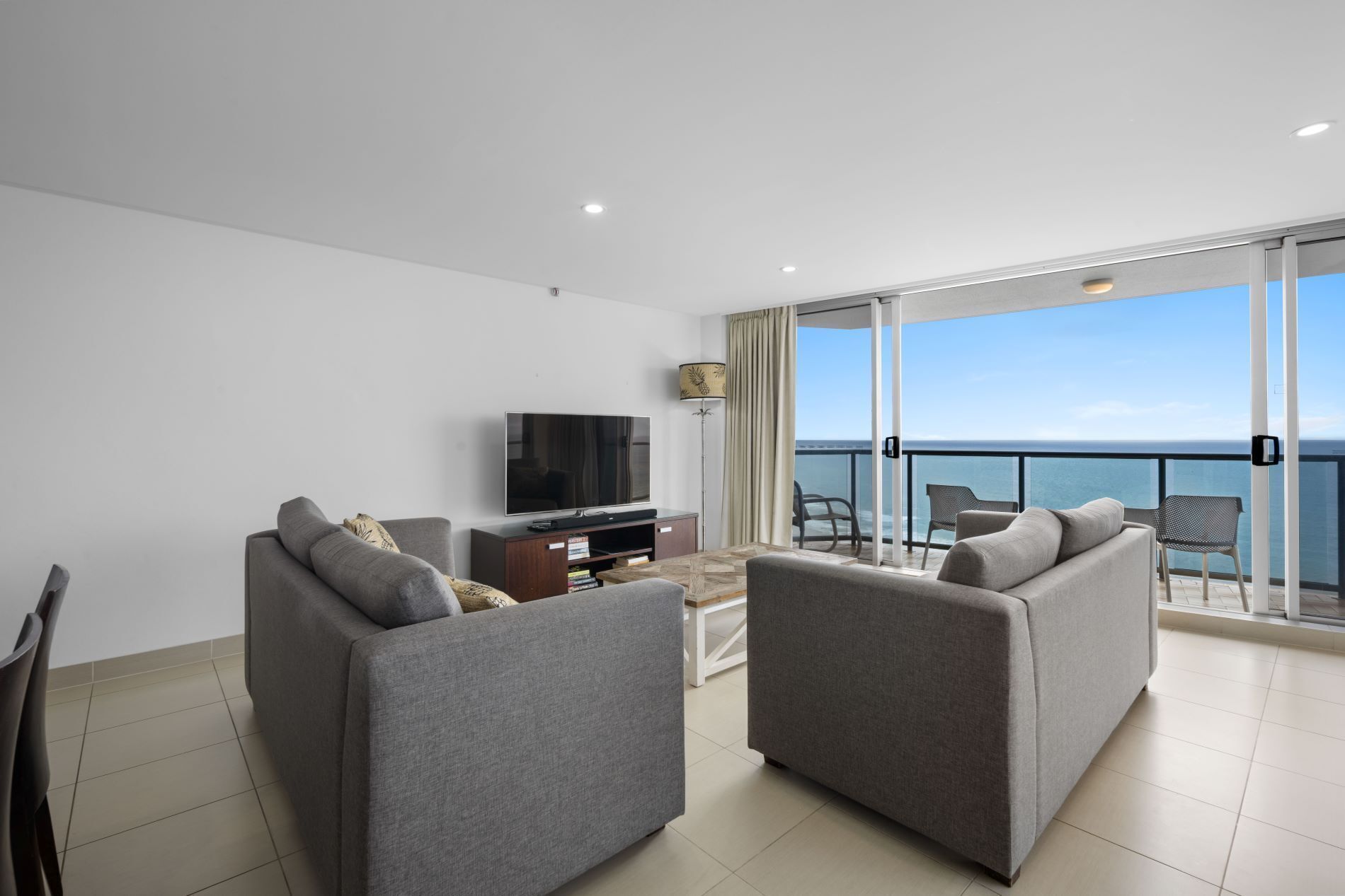 A modern living room with two grey sofas, a flat-screen TV, and a small coffee table. A balcony with a glass door leads to a view of the sea.