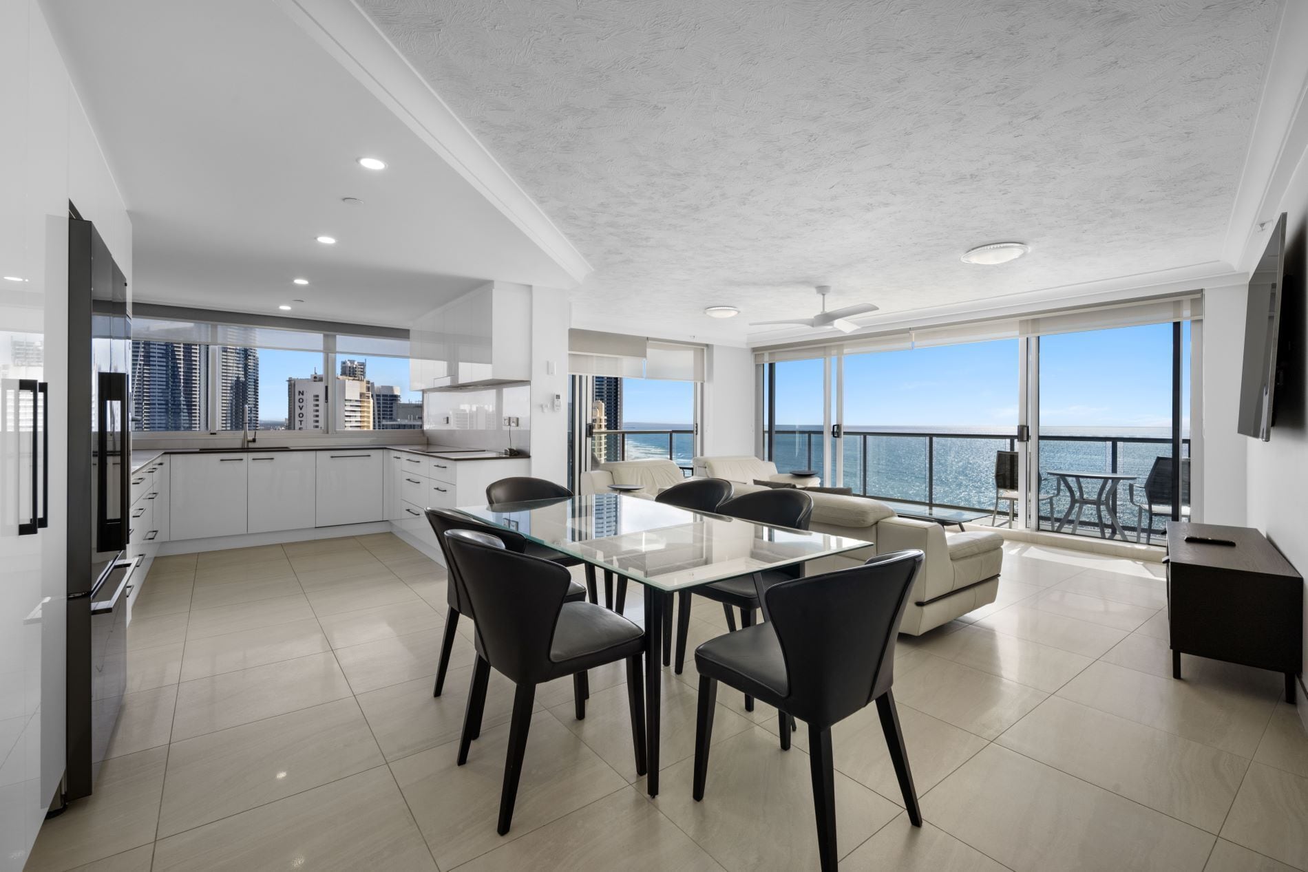 A modern kitchen with white cabinets, a dining area with a glass table, and a balcony with a view of the ocean.