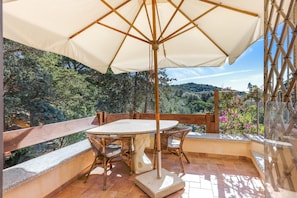 Un patio exterior con una mesa y sillas, barandal de madera y vistas a árboles y colinas.