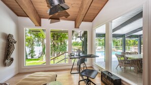 Un'ampia stanza con grandi finestre, soffitto in legno, zona pranzo con mobili in vimini e vista su un'area piscina.