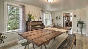 A spacious dining area with a wooden table and benches, a piano, and a view of the kitchen through an open doorway. - Charming Dairy Farm Home in Indiana Amish Country (Shipshewana)