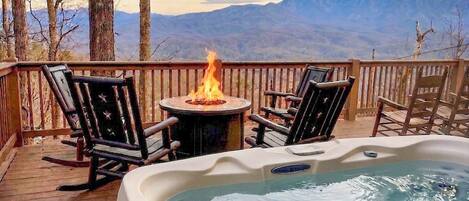 Een hottub op een houten terras met een vuurplaats, omringd door stoelen en een uitzicht op bergen.