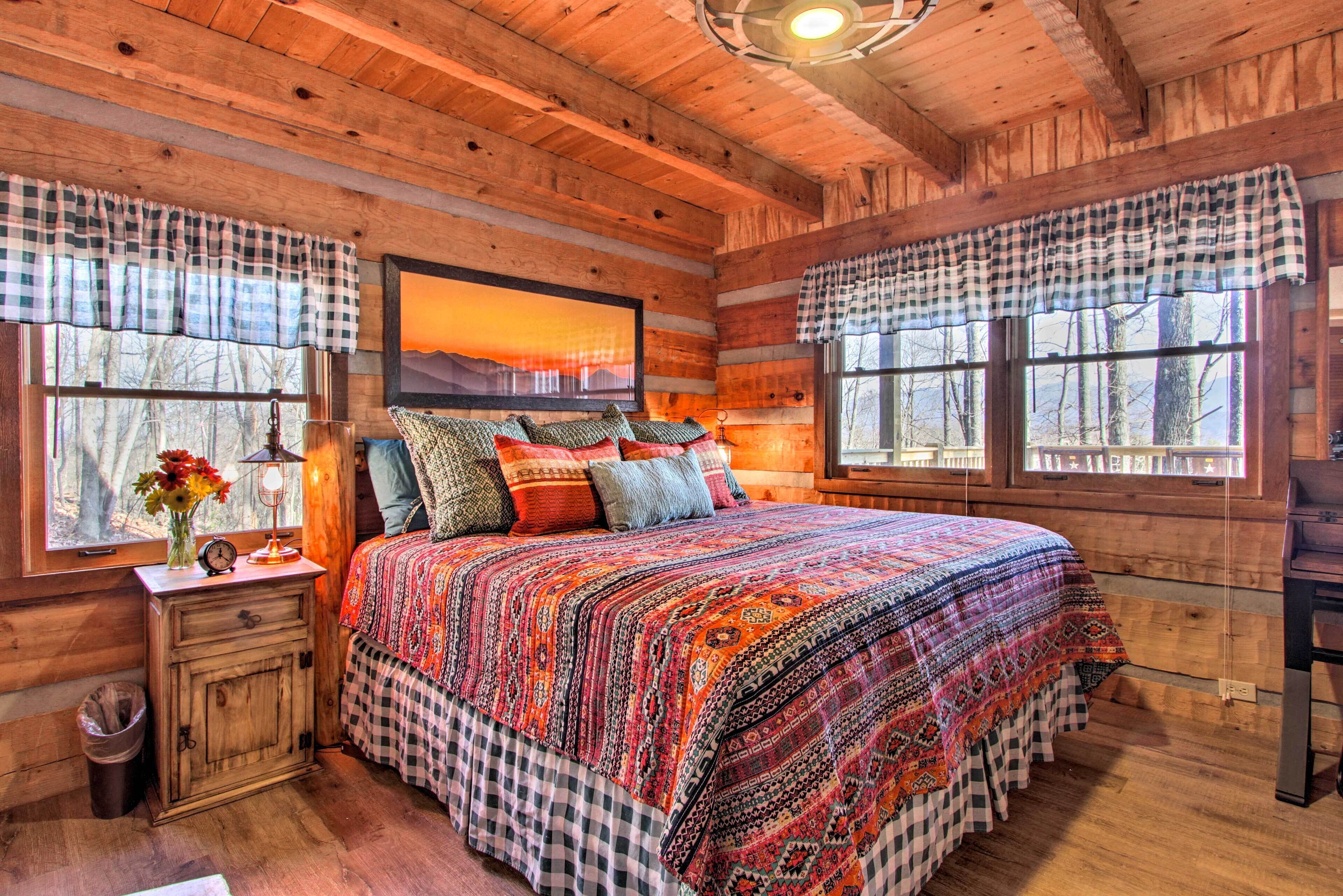 A cozy cabin-style bedroom with a large bed, checkered curtains, a painting of a sunset, and a wooden ceiling.
