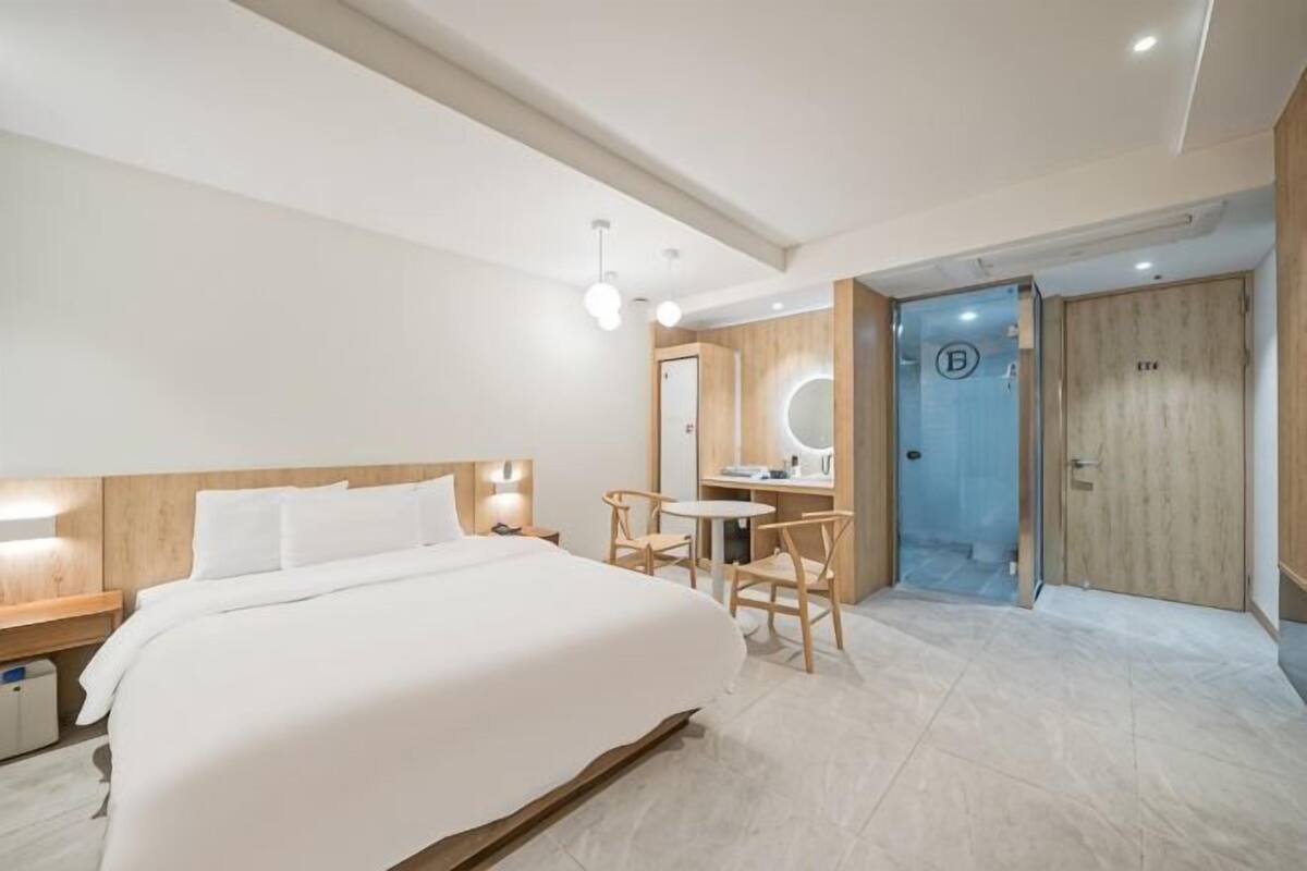 A modern hotel room with a large bed, a desk with a chair, a round mirror, and a wooden door.