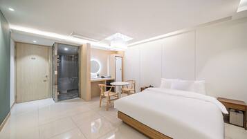 Una habitación de hotel moderna con una cama grande, un escritorio con silla, una mesita redonda y un espejo, además de un cabina de ducha.
