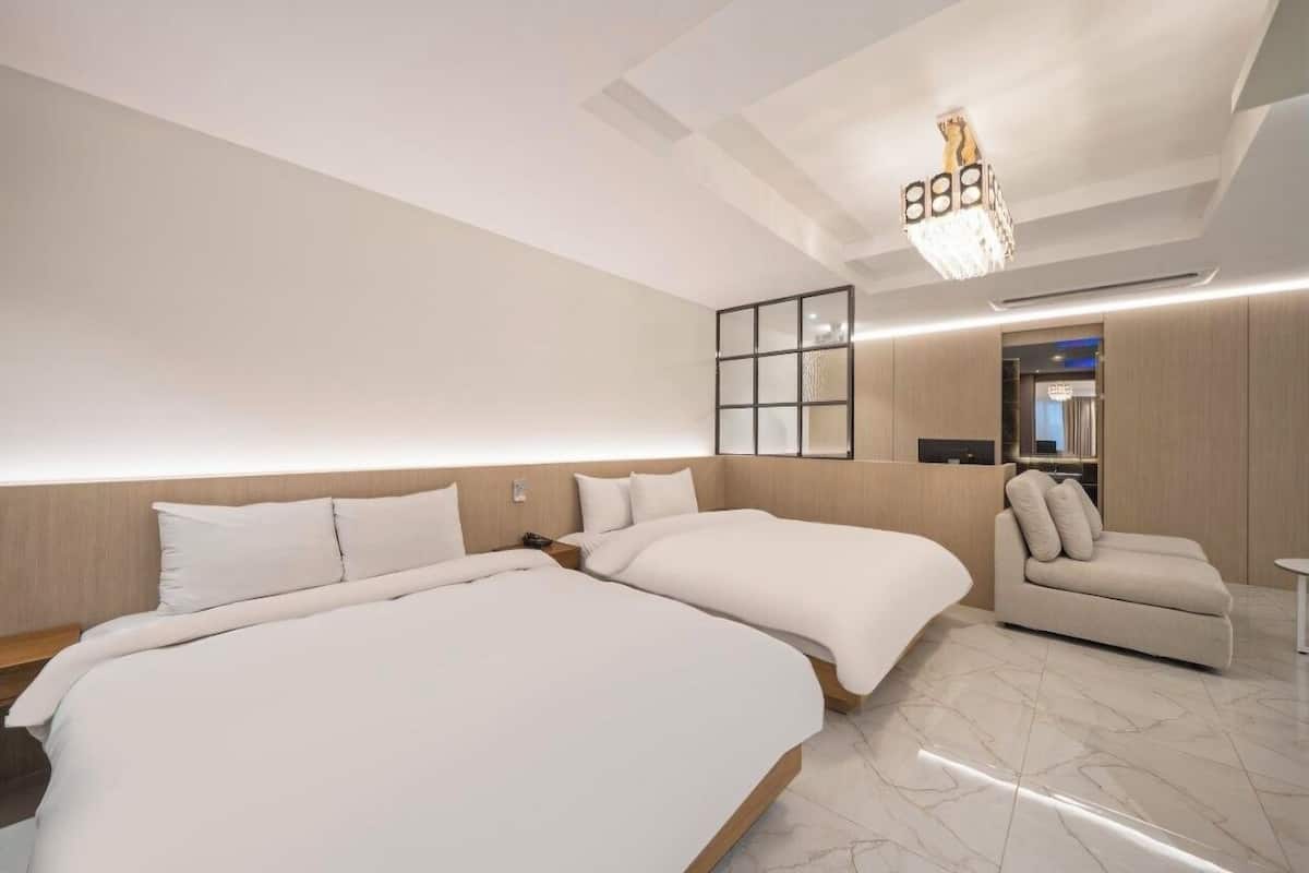 A modern hotel room with two large beds, a small sofa, a desk, and a chandelier.