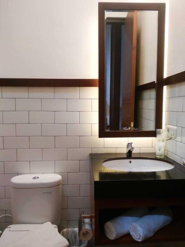 A bathroom with a white toilet, a sink with a mirror above it, and a towel rack with towels. - Are You And I Bed & Breakfast (Bandung)