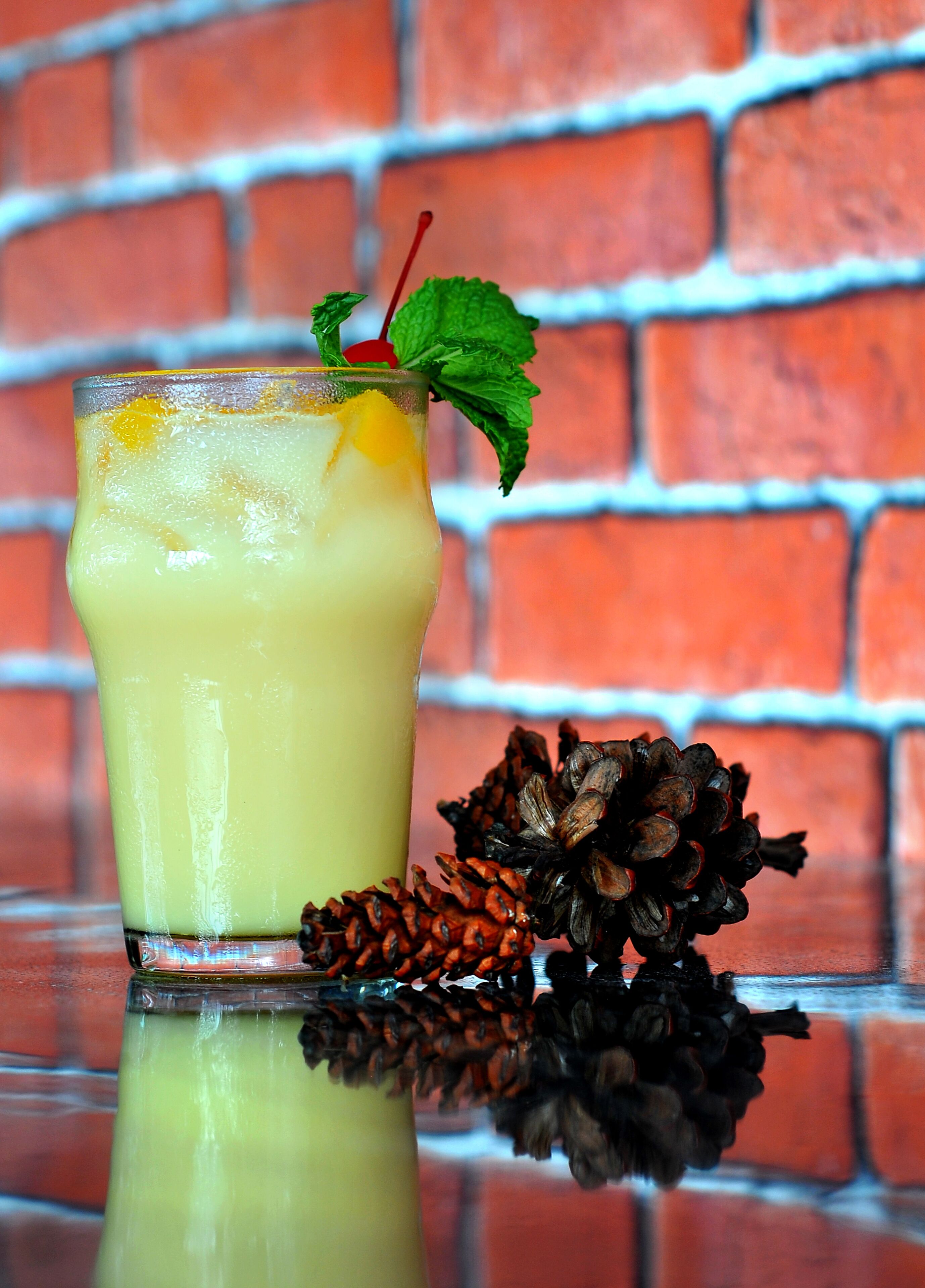 A glass of yellow beverage with a red straw and mint garnish, placed on a reflective surface with pine cones and a brick wall in the background.